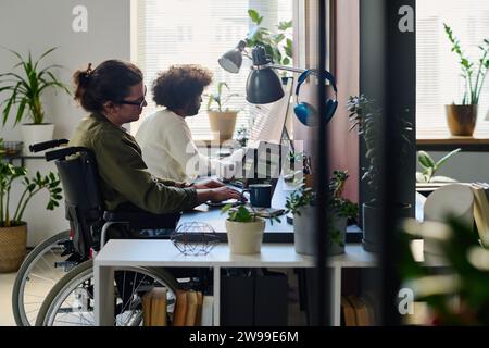 Young businessman in wheelchair looking through AI design task points on laptop screen while sitting by workplace against male colleague Stock Photo