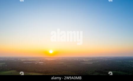 This captivating image showcases the tranquil beauty of a sunrise as it casts a warm glow over a vast landscape. The horizon is adorned with a gradient of orange and blue hues, signifying the break of dawn. The foreground reveals a stretch of lush greenery, possibly farmland or a natural reserve, which is partially shrouded in the soft morning mist. The sky, awash with the light of the rising sun, transitions from a rich blue at the zenith to a golden yellow near the horizon. Serene Sunrise Over a Lush Landscape. High quality photo Stock Photo