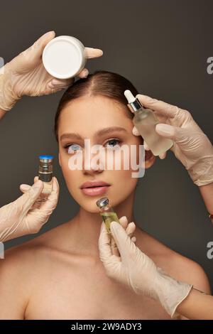 estheticians in medical gloves holding different beauty treatment products near young woman on grey Stock Photo