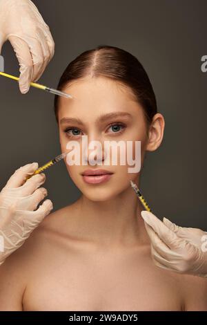 estheticians in medical gloves holding syringes near young woman on grey background, collagen Stock Photo