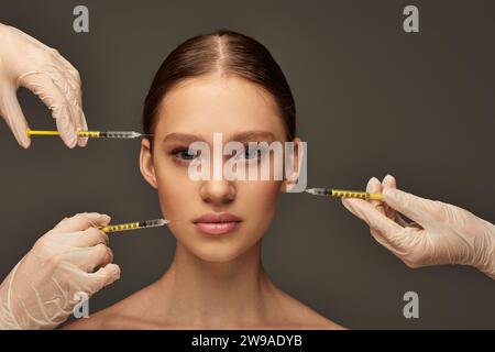 estheticians in medical gloves holding syringes near young woman on grey background, procedure Stock Photo