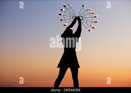 Silhouette of young woman dancing with fire fans against sunset sky in nature, copy space Stock Photo
