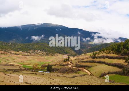 The picturesque Ura Valley in spring with clouds over the mountains, a winding road, farmland, forest and Ura Village, Bumthang District, Bhutan. Stock Photo