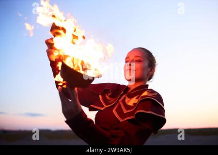 Waist up portrait of smiling young woman dancing with fire bowls outdoors in nature and looking at flames Stock Photo