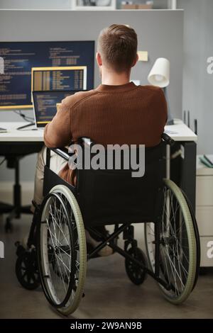 Vertical back view of man with disability writing code while working at desk in office cubicle Stock Photo