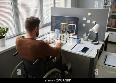 Back view of adult man with disability using wheelchair at office workplace and coding in IT development Stock Photo