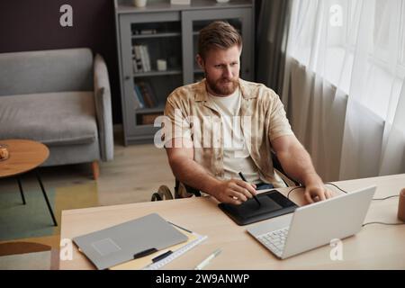 High angle portrait of young bearded man with disability using pen tablet while working as digital artist at home office, copy space Stock Photo