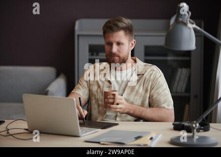 Portrait of young bearded man enjoying cup of coffee while working from home and using pen tablet for editing, copy space Stock Photo