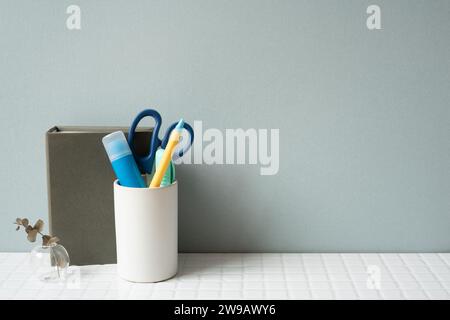 Notebook and holder with glue stick, pen, scissors on white tile desk. gray wall background Stock Photo