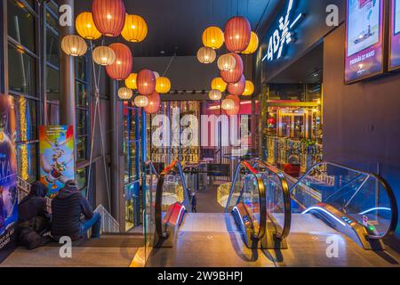 View of couple of young people sitting on steps next to escalator leading into Christmas-decorated lobby of shopping center. Stock Photo