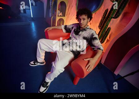Relaxed young man in fashionable outfit sitting in armchair in modern art space looking at camera Stock Photo