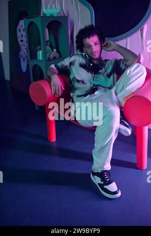 Young relaxed man posing in armchair in studio with vibrant neon lights and looking at camera Stock Photo