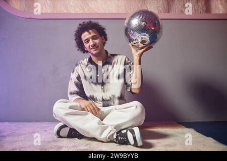 Young relaxed man sitting cross-legged on carpeted floor and holding disco ball looking at camera Stock Photo