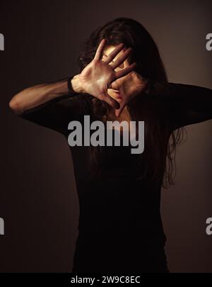 Beauty young witch with long dark hair conjures the hand covering the face on studio dark background in shadow light. Magic concept portrait. Focus on Stock Photo