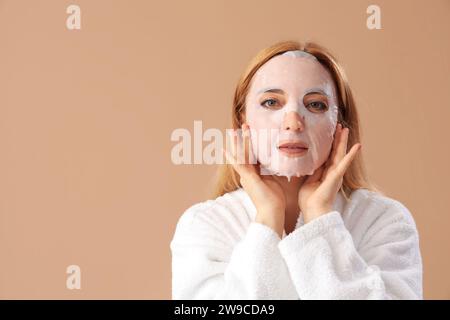 Mature woman in bathrobe with facial sheet mask on beige background, closeup Stock Photo