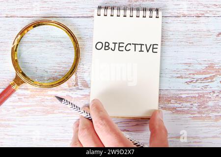 OBJECTIVE written on a blank sheet of a notebook which is touched by a man's hand with a pencil and a magnifying glass is lying next to it Stock Photo