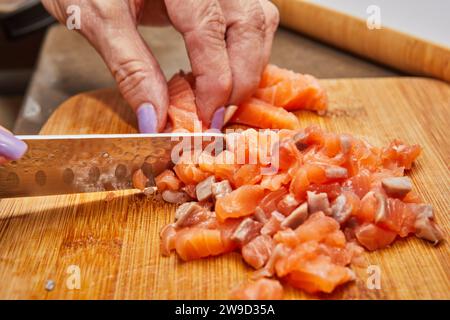 Closeup of hands using sharp knife to slice fresh piece of salmon on wooden cutting board in a well lit and modern kitchen environment. Stock Photo