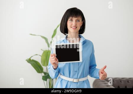 Portrait of young confident modern smiling businesswoman in formal blue shirt shows digital tablet with empty black screen, and looks at camera. Empty Stock Photo
