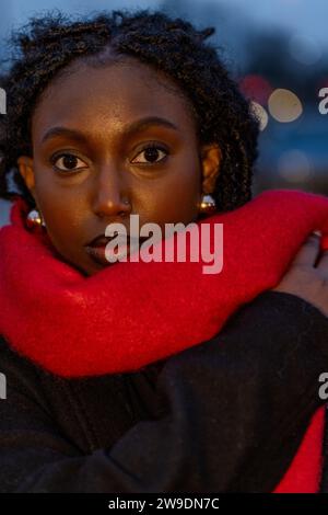The portrait showcases an alluring Black woman wrapped in a vivid crimson scarf, set against the twilight hues of the city. Her captivating eyes are a focal point, drawing the viewer in, while the cool evening light softly illuminates her features, enhancing the rich tones of her skin. The depth of her gaze and the red scarf's contrast against her dark attire add a layer of mystery and sophistication to the composition. Enigmatic Evening in Crimson. High quality photo Stock Photo