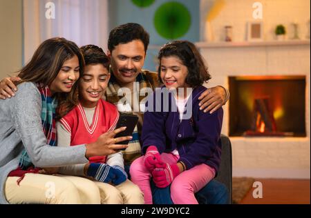 Joyful parents with kids using mobile phone with winter wear at home - concept of social media sharing, technology and weekend holidays Stock Photo