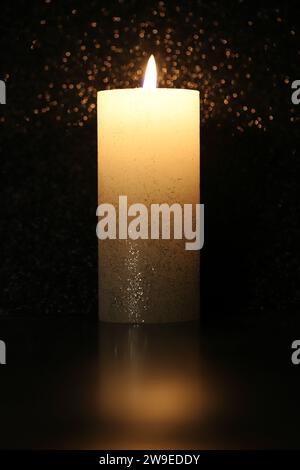 Close-up of a burning white pillar candle with a reflection of candlelight on the floor, black background, side view Stock Photo