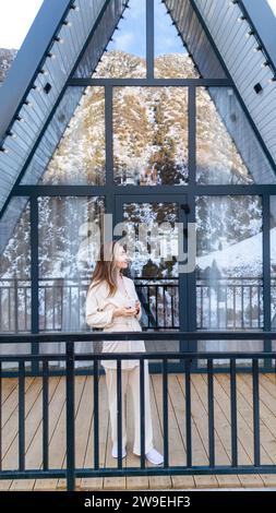 Woman standing and holding cup of tea on the balcony of A Frame cabin in the winter park Stock Photo