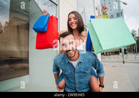 Joyful young man giving a piggyback ride to his girlfriend at store showcase. Happy couple smiling walking flirting at mall carrying a shopping bags. Husband and wife having fun together in a boutique. High quality photo Stock Photo