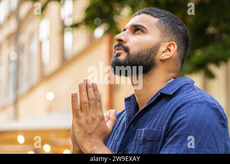 Portrait of bearded religion indian man praying with closed eyes to God asking for blessing, help, forgiveness outdoor. Adult guy clasping hands wishing luck in urban sunny city street. Town lifestyle Stock Photo