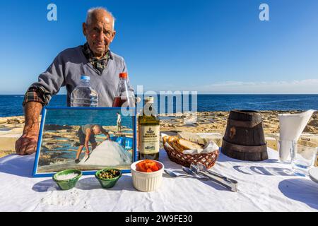 Żaren Darmanin stands at a table set with sea salt, olives, tomatoes, bread and olive oil at the Marsaskala salt pans, Malta, holding a framed photo of himself harvesting salt in summer. Stock Photo