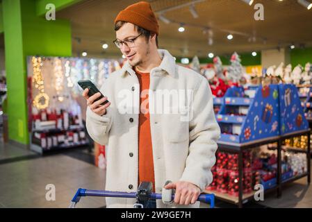 Man in supermarket uses phone. Happy satisfied young caucasian guy in store on background of New Year toys, garlands. Concept of online shopping, internet delivery of everything need to home. Stock Photo