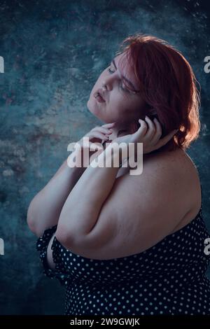 vertical artistic portrait of plus size young latin woman with white skin, with short red hair, standing posing with blue illuminated background Stock Photo