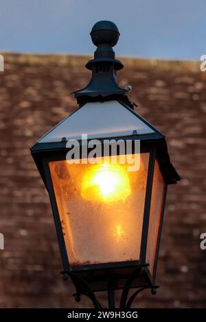 Lit lantern old style outdoor electric metal frame and four glass sides with metal pyramid shaped top church roof in background with some sky portrait Stock Photo