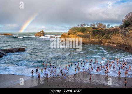 Crowd of swimmers entering the water for the traditional Christmas swim at the Port-Vieux beach with a beautiful rainbow over the Atlantic ocean Stock Photo