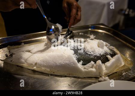 Two spoons lift away the cracked salt crust from a baked fish on a metal tray at Tartarun Fish Restaurant in Marsaxlokk, Malta, revealing the dark skin beneath. The restaurant has been run since 2009 as a family business by brothers James (head chef) and Stephen Schiavone (front of house), evolved from the earlier establishment 'Harbour Lights'. Stock Photo