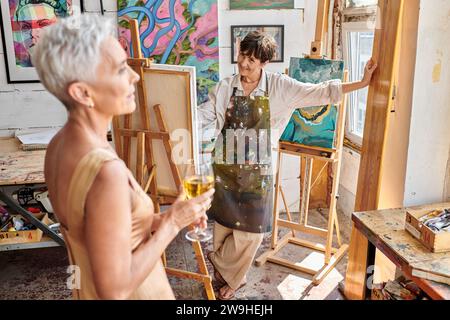 happy woman artist looking at elegant middle aged model posing with wine glass in art workshop Stock Photo