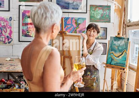 inspired woman artist looking at blurred middle aged model posing with wine glass in art workshop Stock Photo