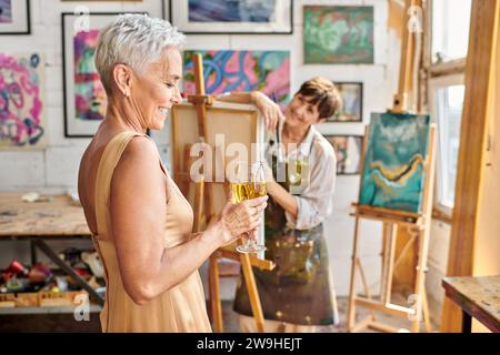 happy and elegant woman posing with wine glass near female artist in workshop, creative process Stock Photo