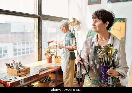 smiling middle aged woman with set of paintbrushes looking away near female friend in art studio Stock Photo