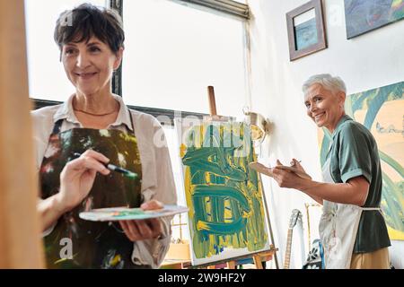 happy mature woman with palette looking at female friend painting art workshop, creative hobby Stock Photo
