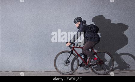 Young guy in casual clothes rides a bicycle against the background of a gray concrete wall. Side view. Urban style. Bicycle commuting concept. Stock Photo