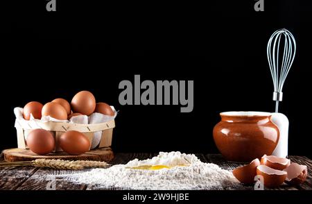 Frozen photo of a broken egg falling in flour. Flour is poured on a wooden board with a texture. In the background is a box with eggs, a whisk and a p Stock Photo
