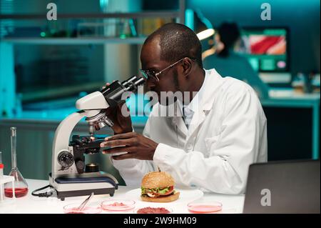 Black researcher attentively looking at piece of lab-grown meat under microscope magnification Stock Photo