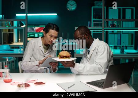 Two scientists analyzing hamburger with lab-grown meat, female researcher taking notes on tablet Stock Photo