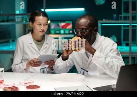 Black scientist smelling hamburger with artificial meat, his asian colleague sitting nearby Stock Photo