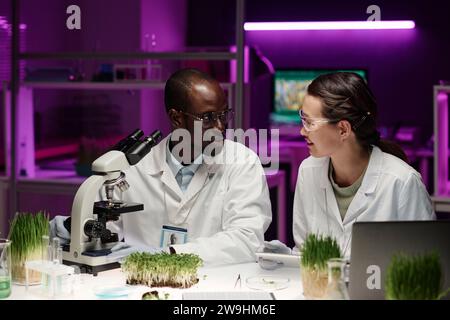 Male and female colleagues discussing scientific work with plants sitting at desk in laboratory Stock Photo