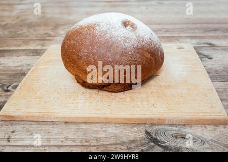 A loaf of freshly baked wheat-rye bread with white topping and a crispy crust on a beech cutting board on an old unpainted wooden tabletop Stock Photo