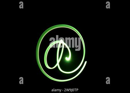 A photograph of an 'at' symbol in vibrant green light in a long exposure photo against a black background. Light painting photography Stock Photo