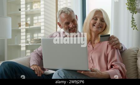 Happy elderly married couple man woman customers hugging making purchase online using laptop talking at home together remote paying with credit card Stock Photo