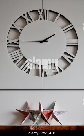 A trio of handmade origami stars on a wooden mantlepiece, under a white wall clock, set against a white background Stock Photo