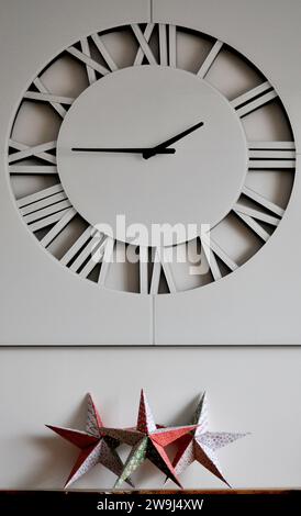 A trio of handmade origami stars on a wooden mantlepiece, under a white wall clock, set against a white background Stock Photo